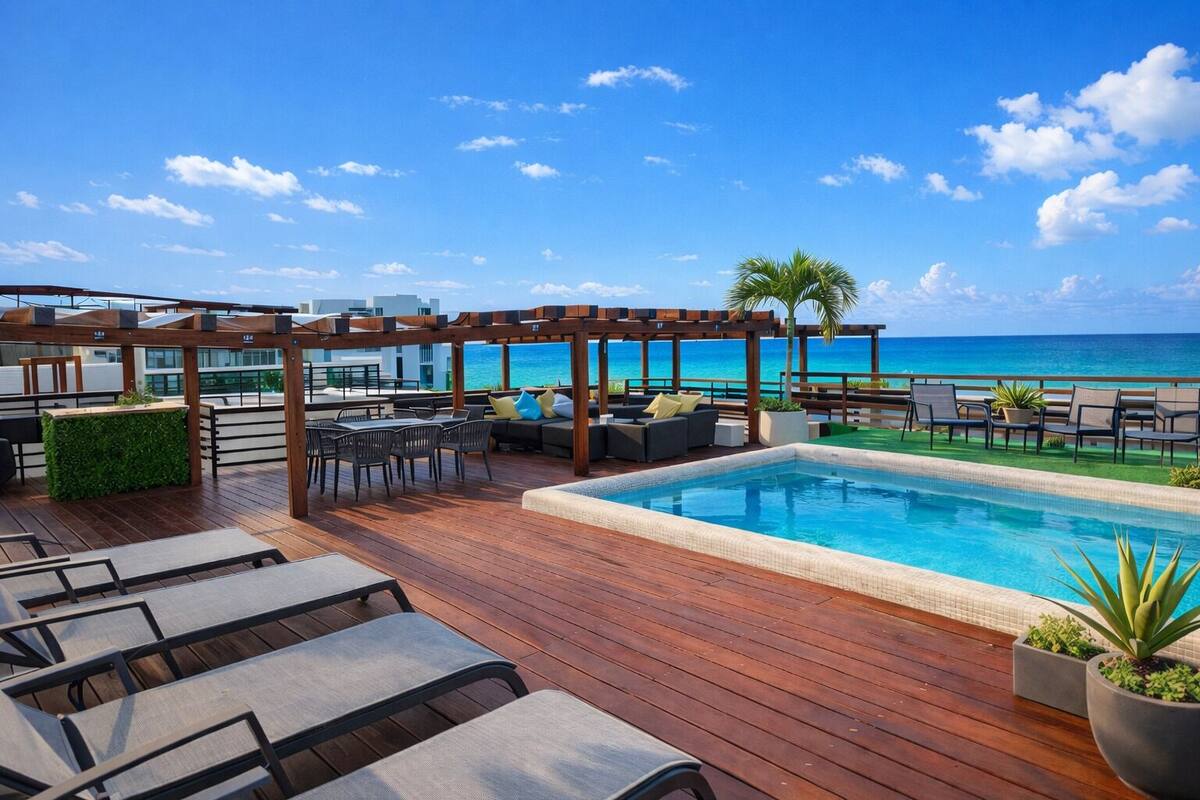 A rooftop pool area with wooden decking, lounge chairs, a small pool, and a view of the ocean.