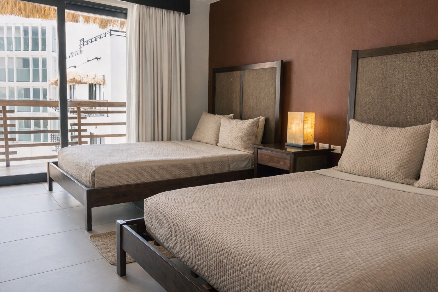 Habitación de hotel con dos camas, un ventanal con cortinas, una mesita de noche con lámpara y vistas a un edificio.