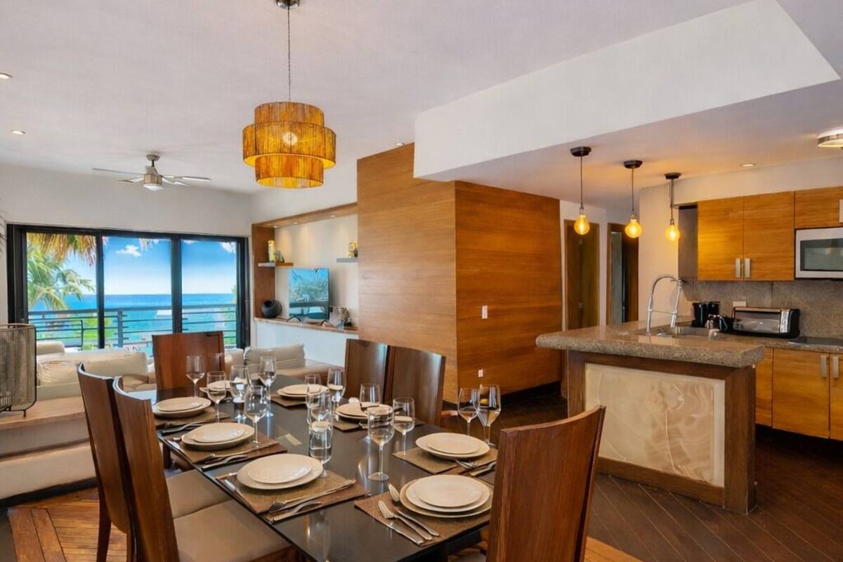 A modern dining area with a wooden table set for six, a kitchen with a granite countertop, and a view of the ocean through large windows.