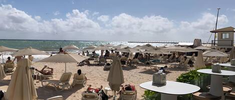 A beachside area with numerous sun loungers and umbrellas, a view of the ocean, and a lifeguard tower in the background.
