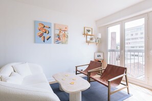 A modern living room with a sofa, a coffee table, and two chairs. There are wall art pieces and a small shelf with decorative items.