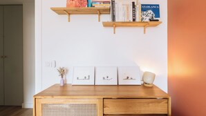 Un escritorio de madera con una cesta tejida, un jarrón con flores secas y tres cuadros enmarcados. Encima del escritorio, hay dos estantes de madera con libros y objetos decorativos.