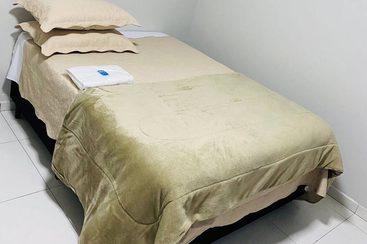 Quarto pequeno com uma cama de solteiro, parede branca simples e chão de azulejos.