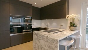 A modern kitchen with dark cabinetry, stainless steel appliances, a marble countertop, and a white orchid plant. - Chelsea Harbour 2-bed Apartment in London (London)