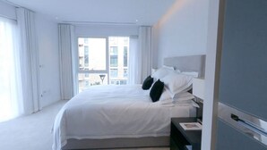 A modern hotel room with a large bed, white bedding, a nightstand, and a view of buildings through the window. - Chelsea Harbour 2-bed Apartment in London (London)