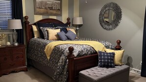 A bedroom with a bed, bedside tables, a nightstand, a ceiling fan, a wall-mounted mirror, and a window with blinds.