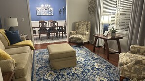 A living room with a blue patterned rug, a sofa, a chair, a table, and a dining area with chairs and a chandelier.