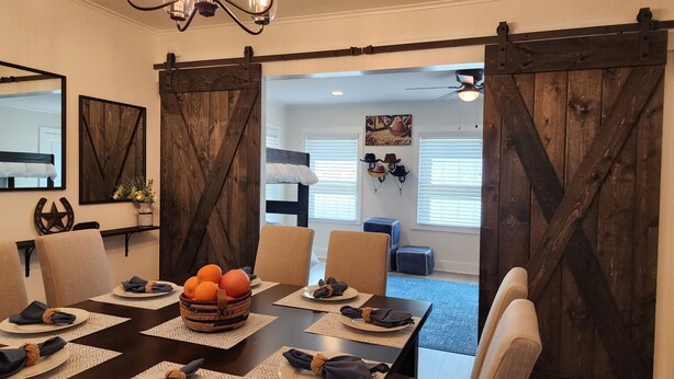A dining area with a dark wooden table set for six, a chandelier, and a view into a bedroom with a bed and a ceiling fan.