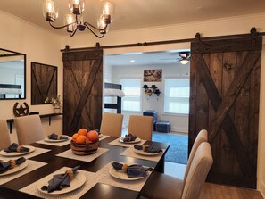 Un coin repas avec une table en bois foncé dressée pour six personnes, un lustre et une vue sur une chambre à coucher avec un lit et un ventilateur de plafond.