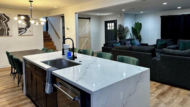 A modern kitchen with a marble countertop, a sink, and green chairs. There is a dark-colored sofa and a staircase in the background. - 🦚The Peacock Inn LV - 8 Bdr 4 Bth 14 Beds, Hi Speed WiFi (North Las Vegas)