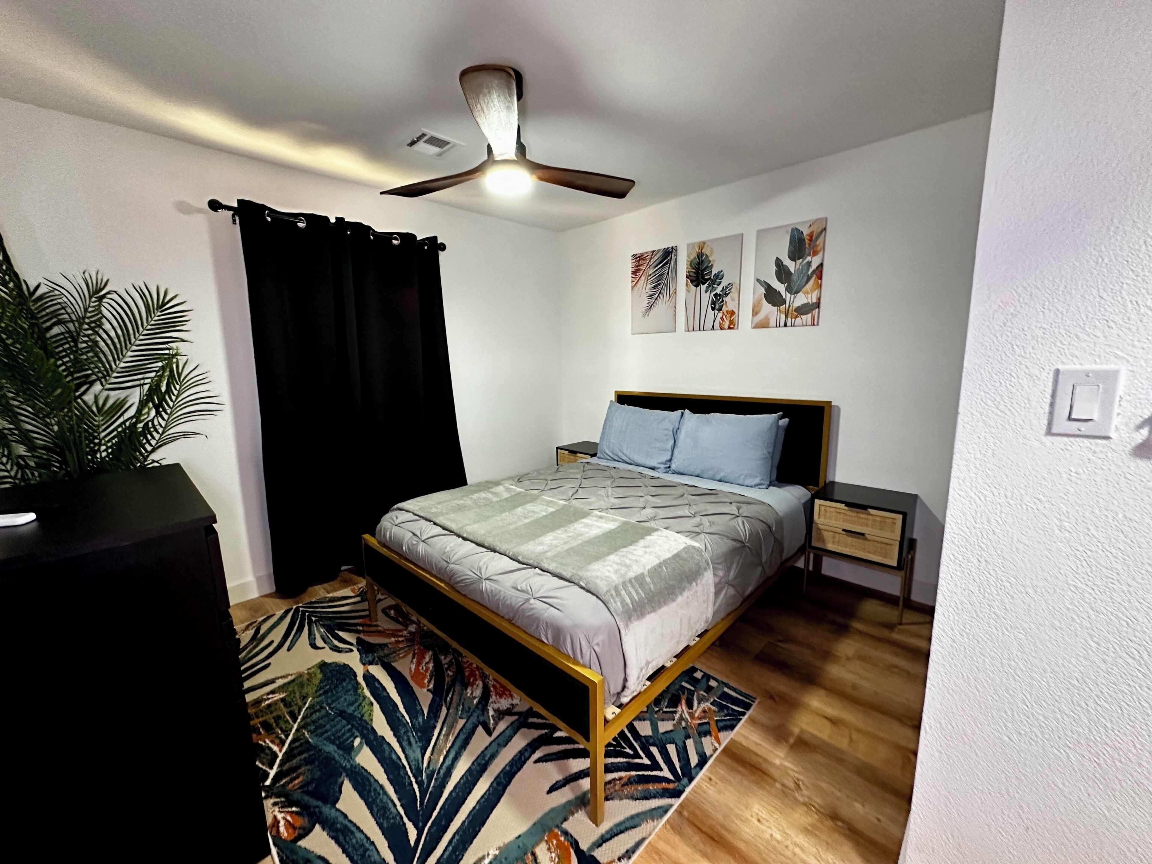 Un dormitorio con cama, ventilador de techo, mesita de noche, interruptor de luz en la pared y una planta en maceta.