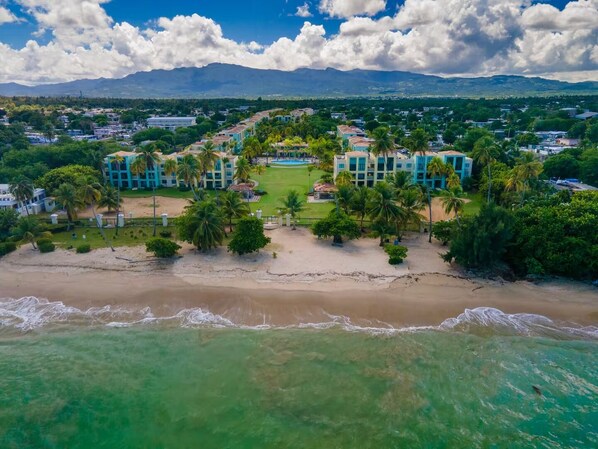 An aerial view of a coastal resort with beachfront villas, palm trees, and a mountain backdrop.