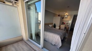 Een hotelkamer met een schuifdeur naar een balkon, een bed met een grijze dekbedovertrek, een klein bureau met stoel, en een kitchenette.