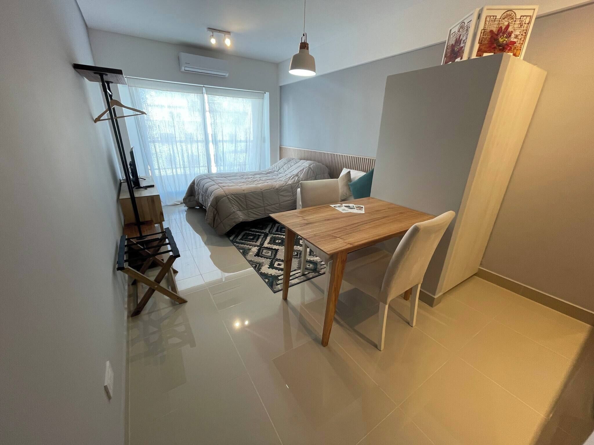 A compact bedroom with a bed, a small table, and a chair. There is a window with curtains, a floor lamp, and a radiator.