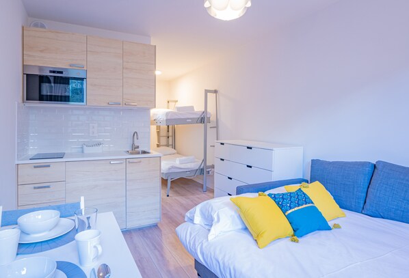 Quarto de hotel moderno com cozinha compacta, cama com almofadas azuis e amarelas, e mesa de cabeceira com xícara e pires.