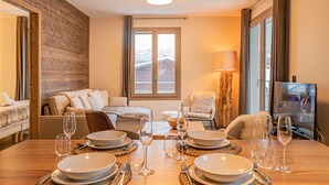 Uma sala de jantar com uma mesa de madeira posta para quatro pessoas, um sofá e uma televisão ao fundo.