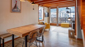 Une table à manger en bois, des chaises, un canapé et un petit tabouret dans une pièce dotée de poutres en bois et de grandes fenêtres.