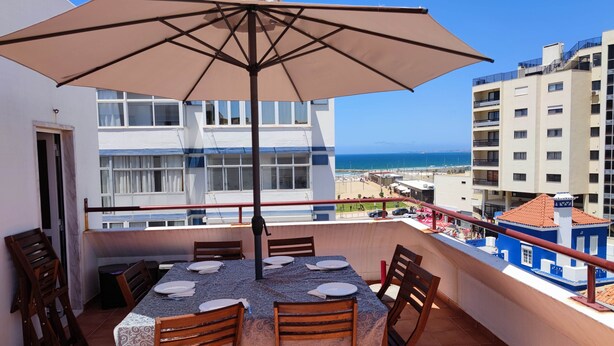 A balcony with a table set for four, an umbrella, and a view of the sea and buildings. - Amazing Terrace Costa da Caparica (Costa da Caparica)