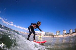 Iemand surft op een rood board in de oceaan, met een strand en gebouwen op de achtergrond.