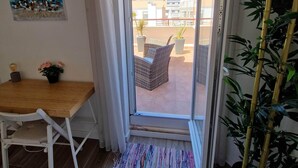 A room with a wooden table and white chair, a sliding glass door leading to an outdoor area with wicker furniture and potted plants. - Amazing Terrace Costa da Caparica (Costa da Caparica)