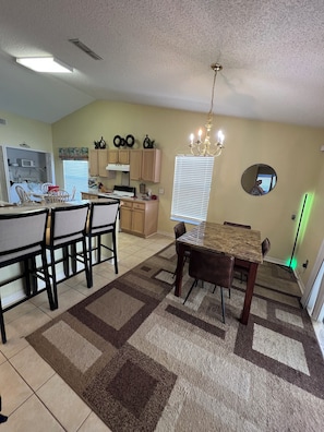 A dining area with a table and chairs, a kitchen in the background, and a chandelier.