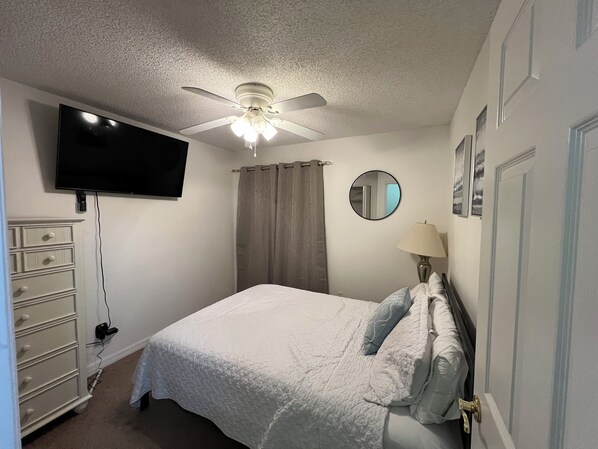A bedroom with a bed, a ceiling fan, a TV, a dresser, a lamp, and a mirror.