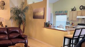 A living room with a brown leather sofa, a coffee table, a large potted plant, and a kitchen in the background. - Near Disney & Universal Pool + Spa Family 4BR Private Pool, Spa Fenced Yard (Kissimmee)
