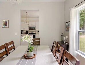 A dining area with a table, chairs, and a potted orchid. There is a kitchen in the background with cabinets and appliances.