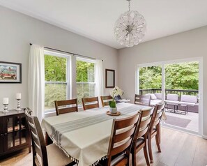 A dining area with a table set for six, surrounded by chairs, a chandelier, and a view of greenery through large windows.