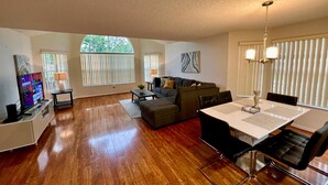 A spacious living room with a wooden floor, a dining area with a white table and black chairs, a gray sofa, and a TV on a stand. - Grand Family Deluxe 3BD Condo Near Disney Universal 22 (Kissimmee)