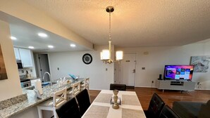 A modern kitchen with granite countertops, a dining area with a white table and black chairs, a living room with a TV and a couch, and a hallway with a clock. - Grand Family Deluxe 3BD Condo Near Disney Universal 22 (Kissimmee)