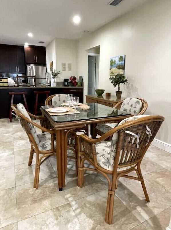 A dining area with a glass-top table surrounded by wicker chairs, a kitchen in the background, and a painting on the wall. - Early Bird Summer Special Weekly Discounts Hibiscus House (Vero Beach)