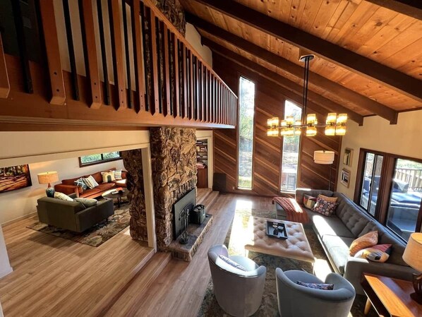 A spacious living area with a stone fireplace, wooden floors, and a high ceiling.