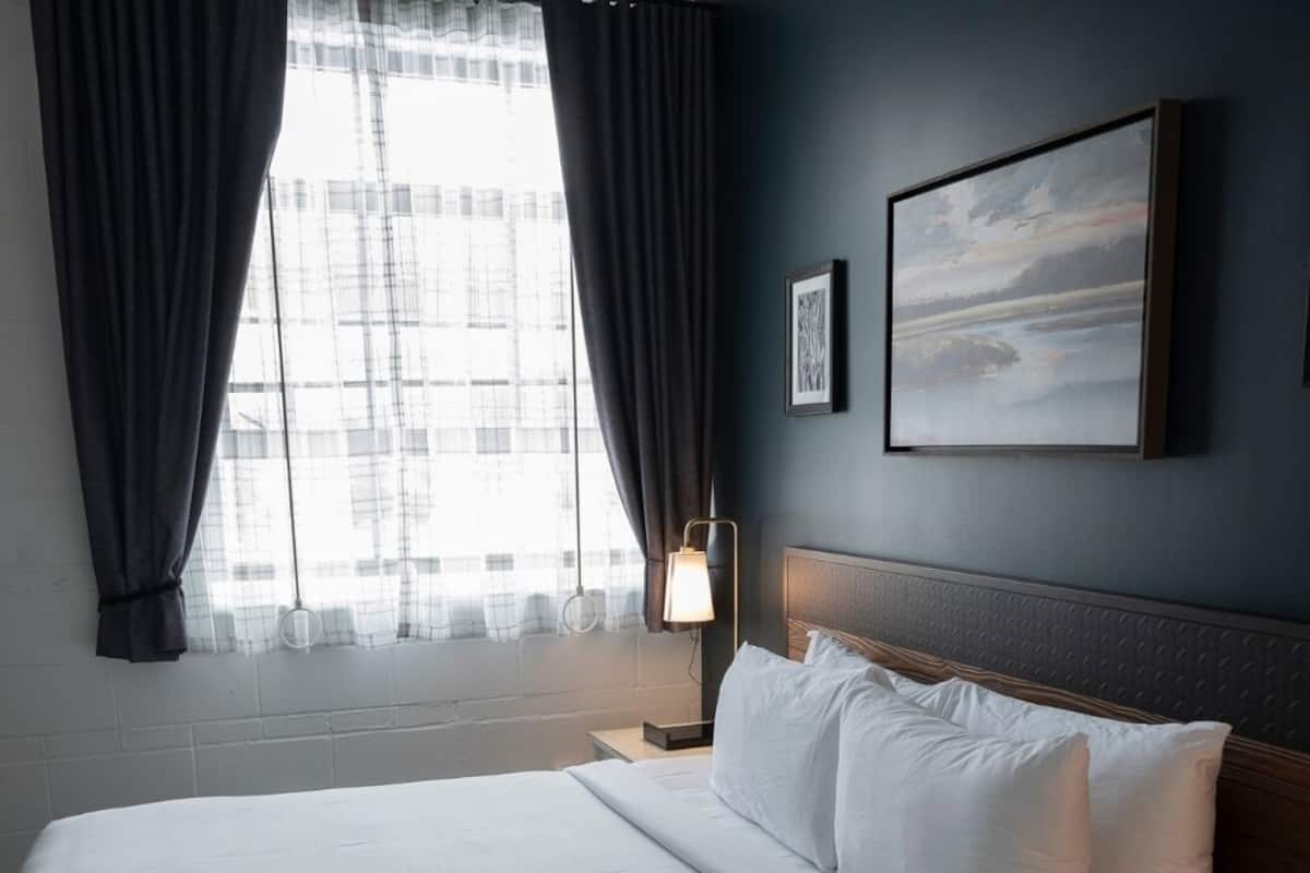 Quarto de hotel com uma cama grande, uma mesinha de cabeceira com abajur, um quadro na parede e uma janela com cortinas.