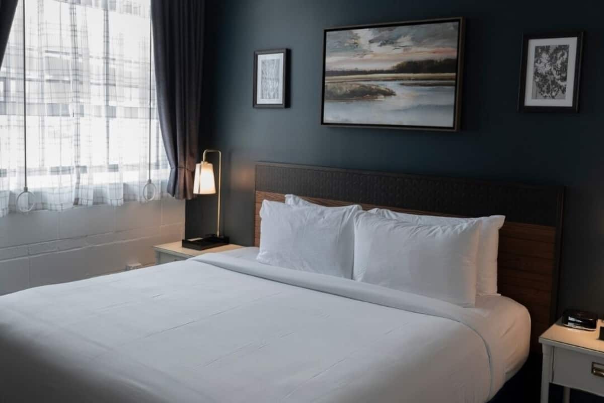 Quarto de hotel com uma cama grande, uma mesa de cabeceira com abajur e vista para um edifício pela janela.