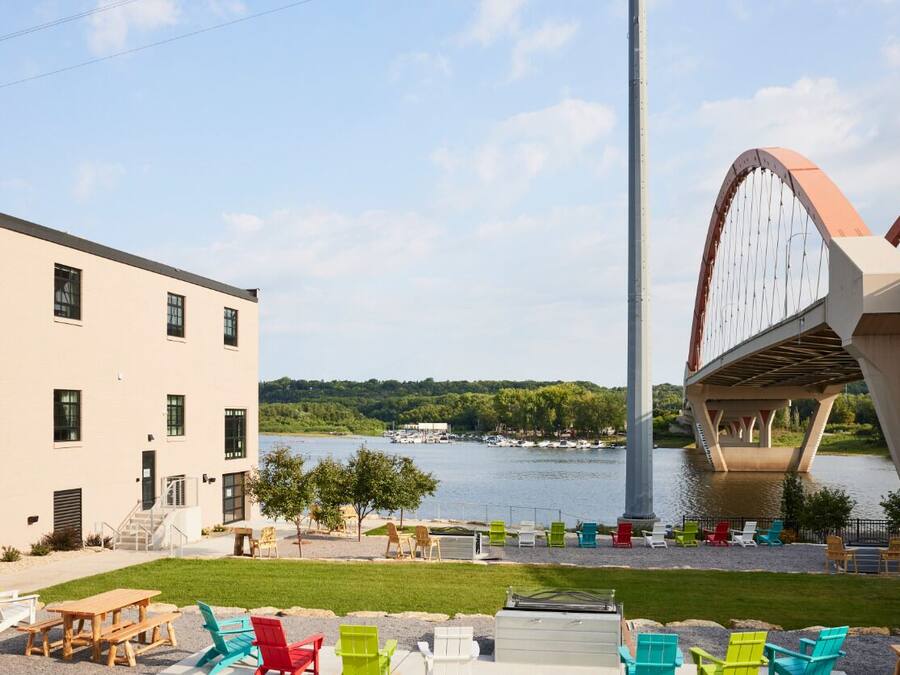 A riverside outdoor seating area with picnic tables and colorful chairs, a modern building with multiple windows, and a distinctive bridge structure in the background.
