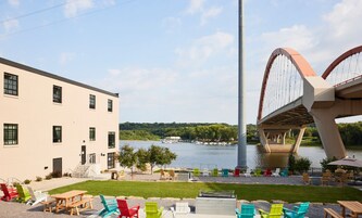 Un área al aire libre junto al río con mesas de picnic y sillas coloridas, un edificio moderno con amplias ventanas, y una estructura de puente distintiva al fondo.