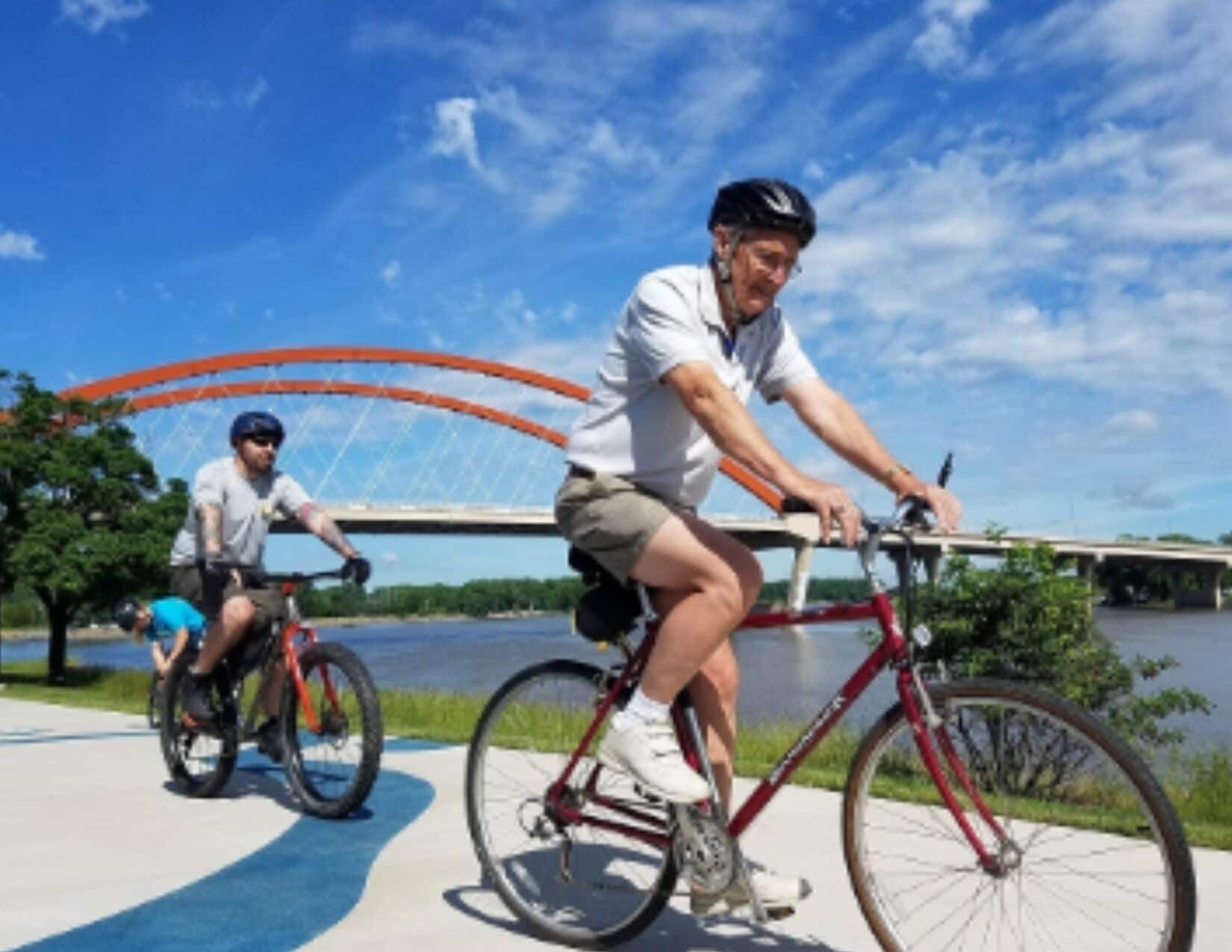 Two cyclists on a paved path with a bridge in the background.