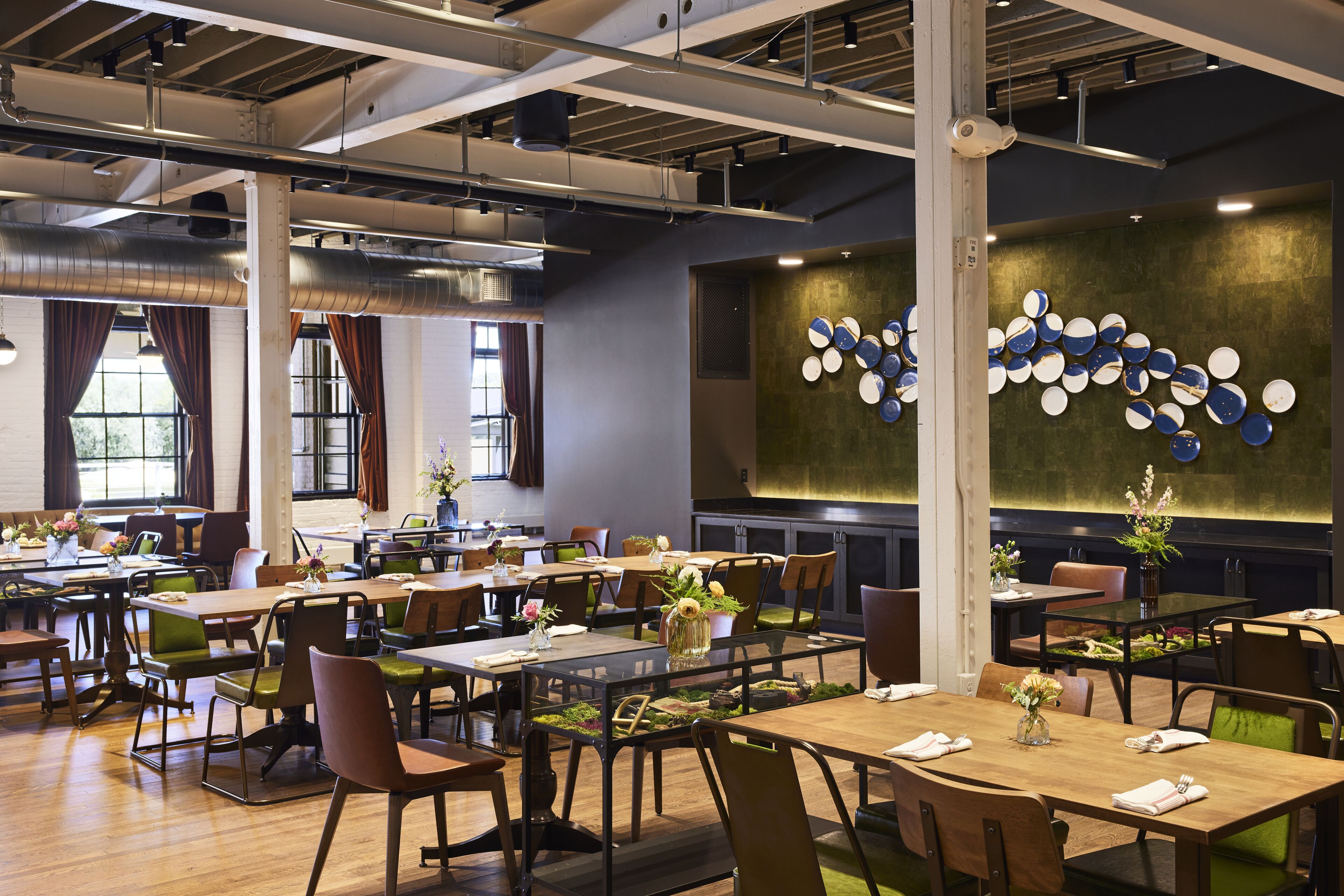 A spacious dining area with multiple tables and chairs, large windows with curtains, and a wall with circular decorative plates.