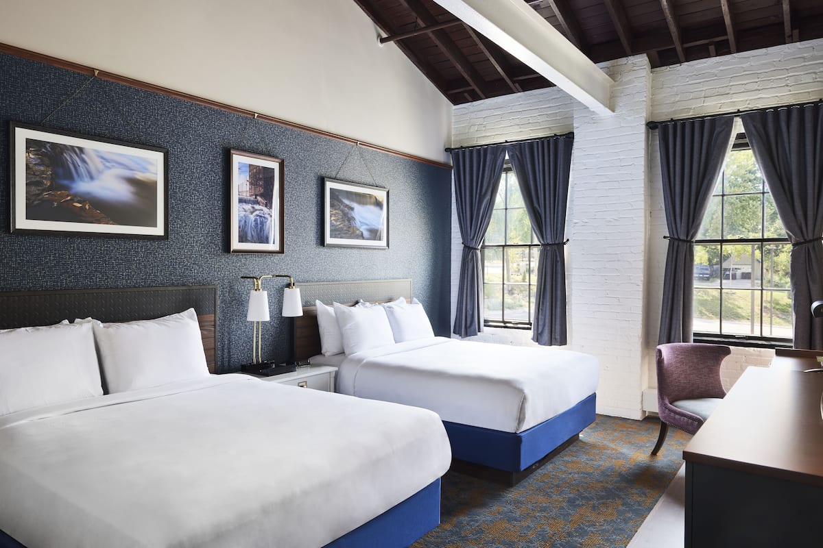 Quarto de hotel com duas camas, uma escrivaninha, uma cadeira e grandes janelas com cortinas.