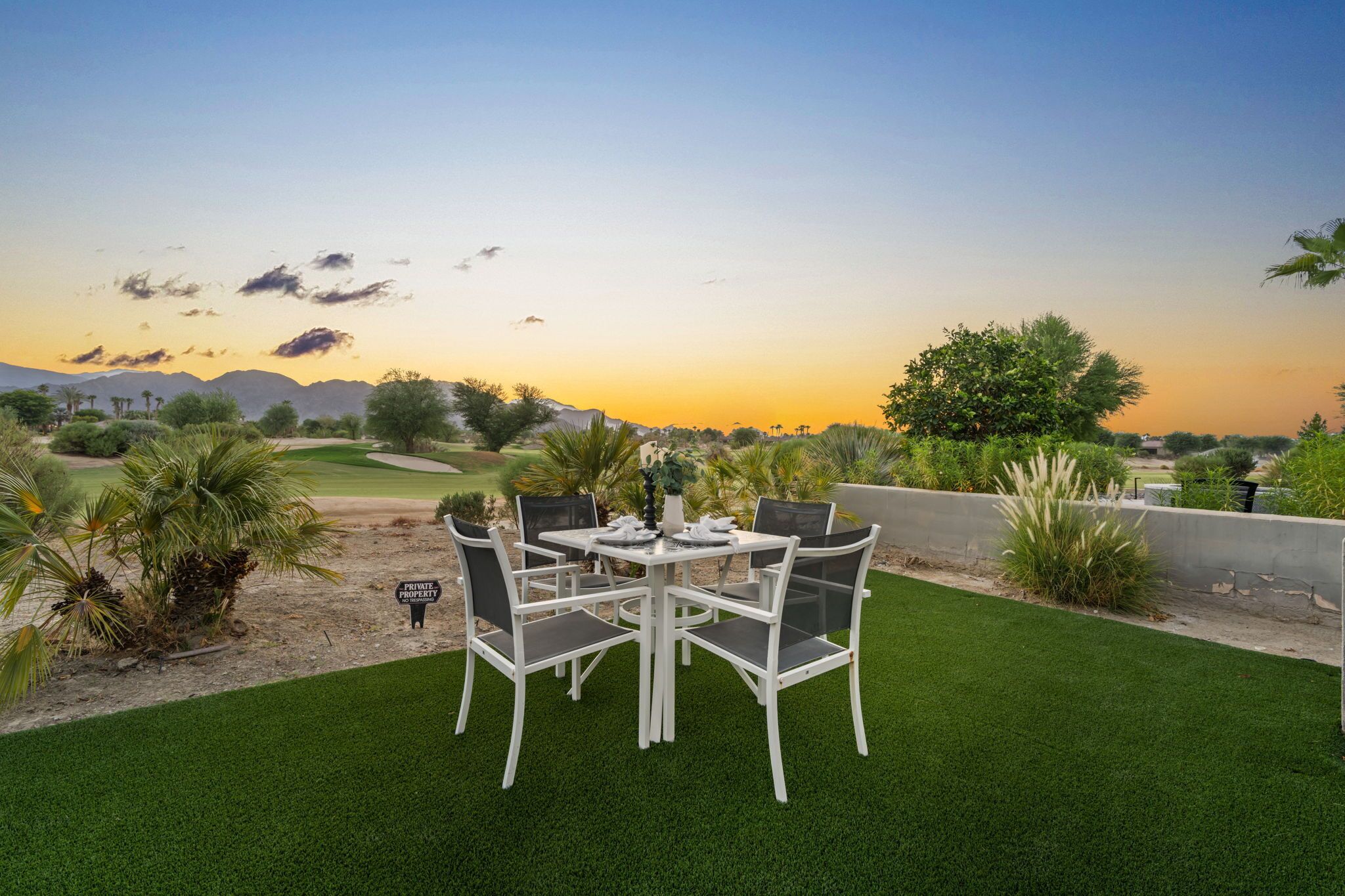 A dining area with a table and chairs, surrounded by artificial grass and plants, with a view of mountains and a sunset in the background.