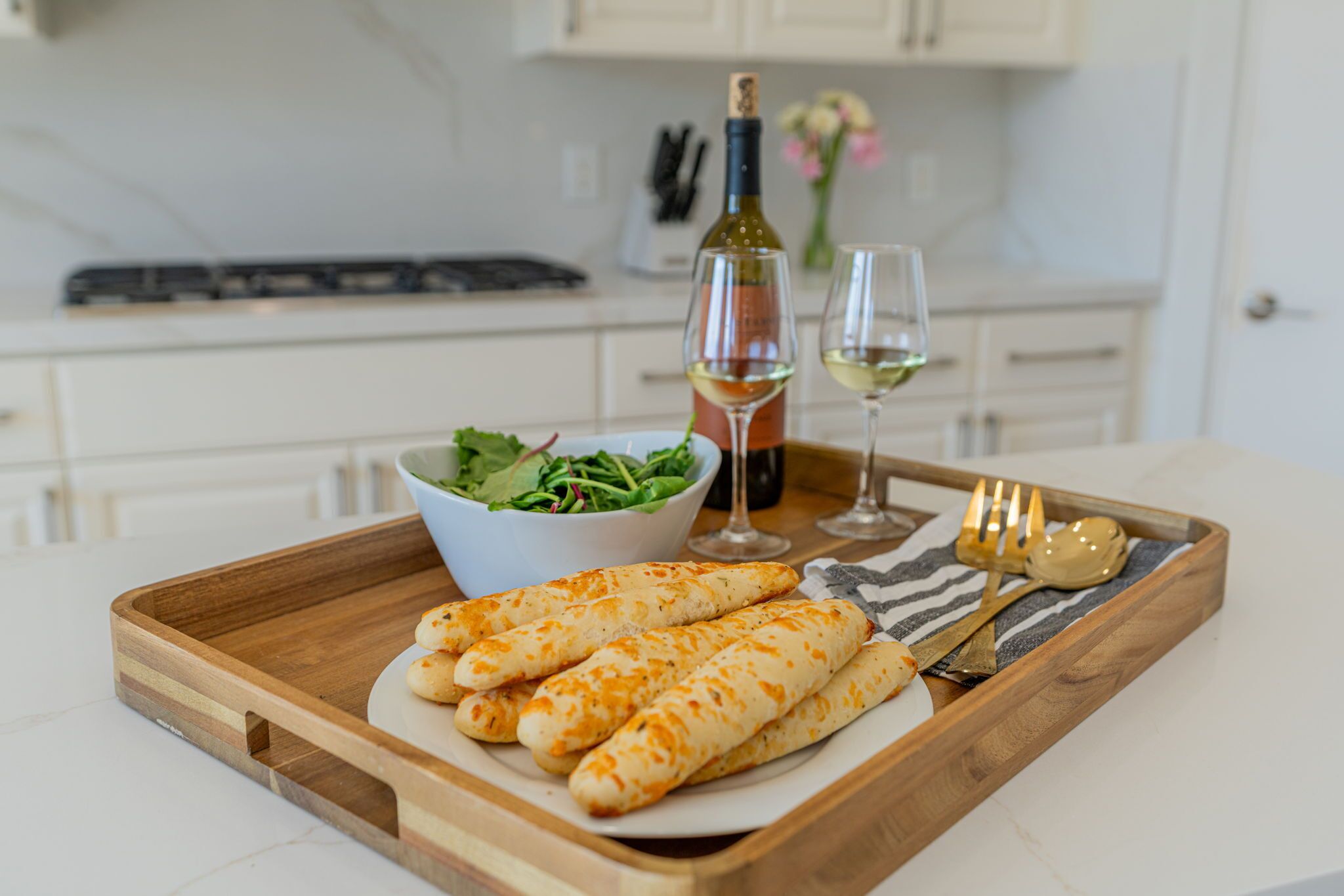 A wooden tray with a plate of cheese sticks, a bowl of salad, a wine bottle, and glasses on a kitchen counter.