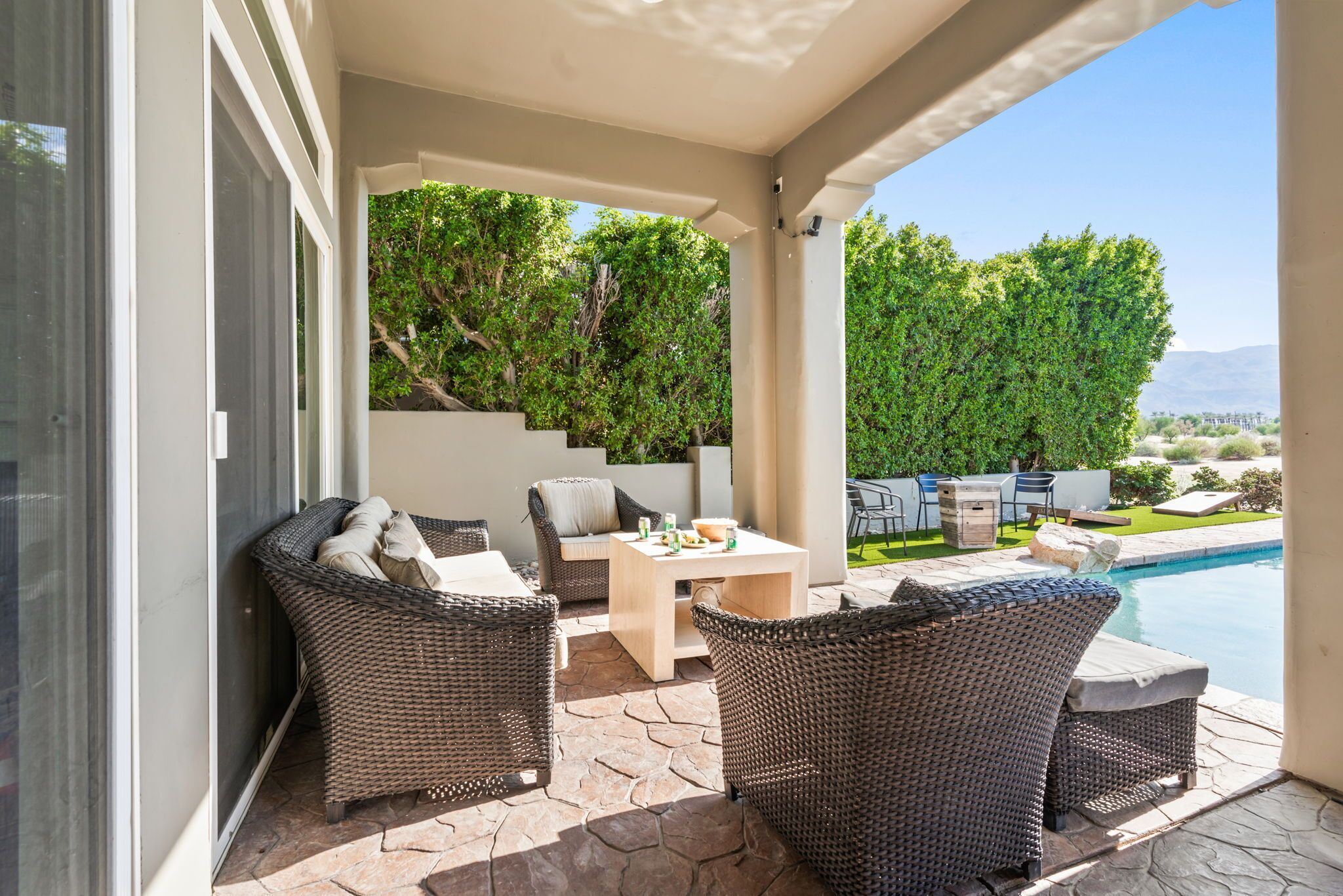 A covered patio with wicker furniture, a table, and a view of a swimming pool and greenery.