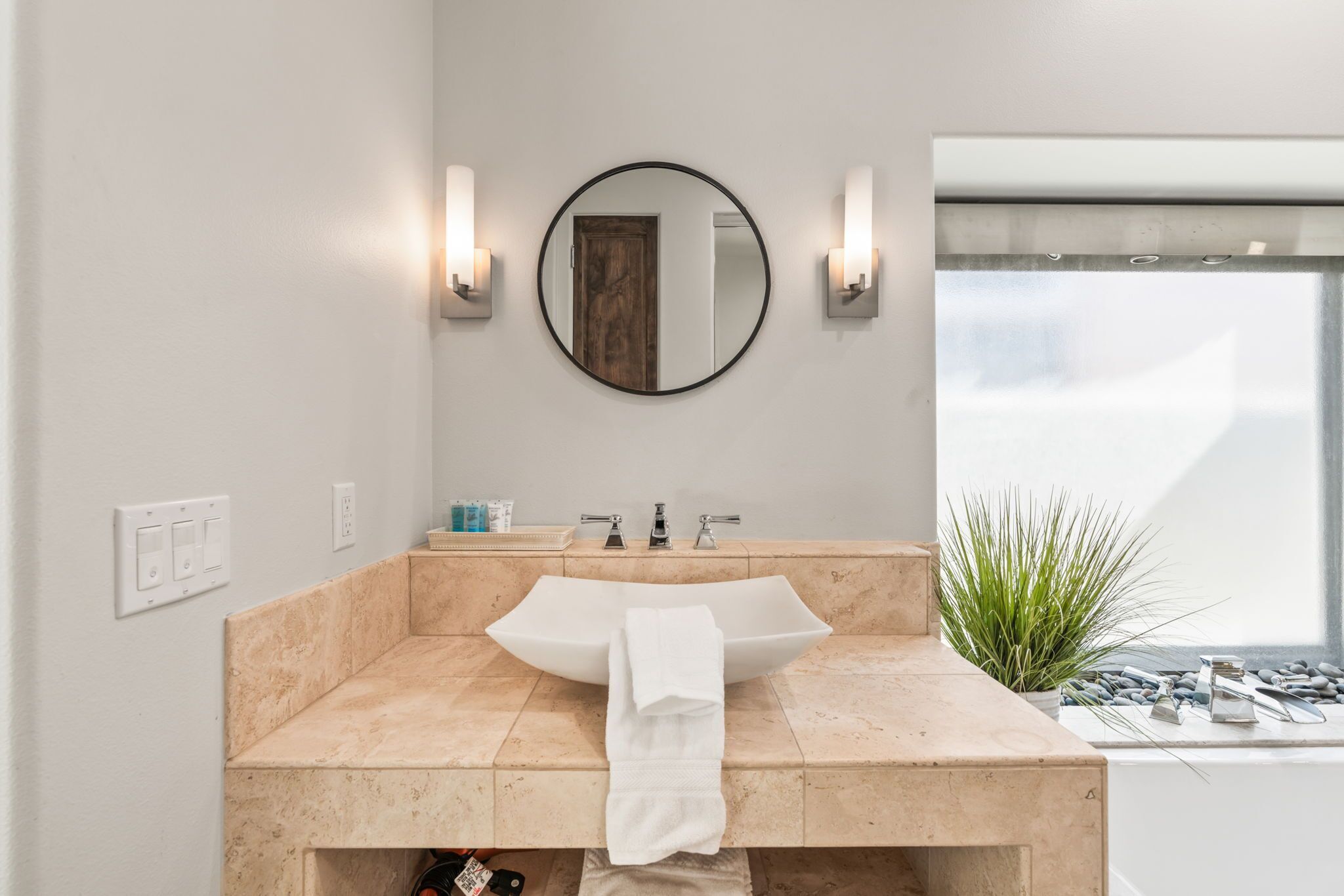 A bathroom with a beige marble sink, a round mirror, and two wall-mounted lights.