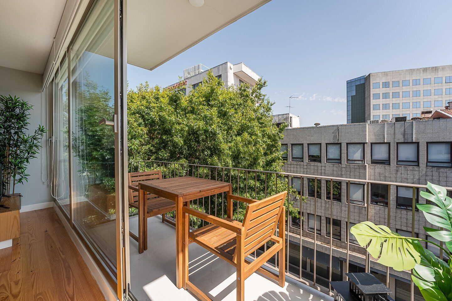 A balcony with a wooden table and chairs, a potted plant, and a view of a modern building and trees.
