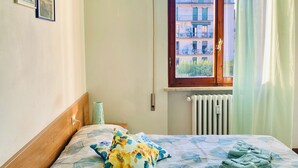 A neatly made bed with a floral bedspread, a bedside table with a tissue box and a bottle, and a window with a view of buildings. - Comfortable two-room apartment near Santa Maria Novella station (Firenze)