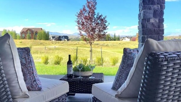 Pátio com móveis de vime e uma mesa com garrafa e copo, com vista para um campo gramado e um edifício ao longe.