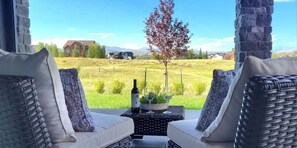A patio with wicker furniture and a table with a bottle and glass, overlooking a grassy field and a building in the distance.