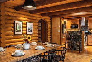 L’intérieur d’une cabane en rondins présente un coin repas avec une table en bois dressée pour quatre personnes, entourée de chaises noires. Un grand vase blanc rempli de fleurs est posé sur la table.