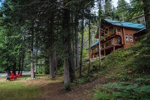Une cabane en rondins dotée d’une terrasse, entourée d’une forêt dense et bordée par une rivière.